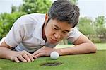 Mid adult man lying down in a golf course trying to blow the ball into the hole