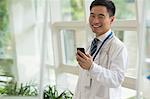 Smiling doctor using his phone in the hospital lobby, looking at camera, glass doors