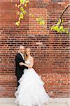 Portrait of bride and groom standing outdoors in front of brick wall, looking at camera and smiling, Canada