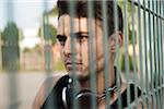 Close-up portrait of young man outdoors, standing behind steel fence with headphones around neck, Germany