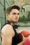 Close-up portrait of young man sitting on ground with basketball, wearing headphones around neck at fenced-in basketball court, Germany