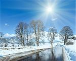 Spring sunshine and road through the alpine village in Austria with reflection of trees in thawing snow.