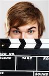 Portrait of a young man peeking behind a clapboard, over a gray background