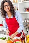 A beautiful happy young woman or girl wearing glasses & a red apron cutting & preparing fresh vegetable salad food in her kitchen at home