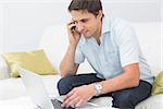 Smiling young man using laptop and cellphone in the living room at home