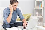 Smiling young man using cellphone and laptop in the living room at home