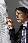 Side view of a serious young businessman peeking through blinds in the office