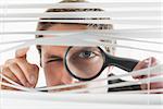 Close-up portrait of a young businessman peeking through blinds with magnifying glass in the office