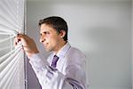 Side view of a young businessman peeking through blinds in the office