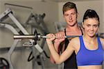 Portrait of a young male trainer helping young fit woman to lift the barbell in the gym