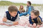 Happy siblings lying with parents sitting in background at the beach