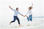 Full length of a cheerful young couple holding hands and jumping at the beach