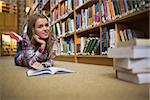 Pretty cheerful student lying on library floor reading book in college