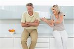 Tired man sitting with arms crossed as woman argue in the kitchen at home