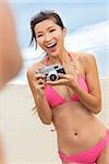 A beautiful Asian Chinese girl or young woman wearing pink bikini at a beach looking happy taking pictures or photographs with a retro digital camera