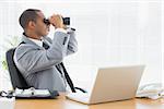 Side view of a young businessman looking through binoculars at office desk