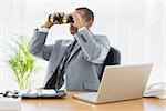 Serious young businessman looking through binoculars at office desk