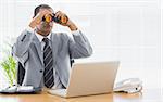 Serious young businessman looking through binoculars in front of laptop at office desk