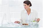 Concentrated young businesswoman using computer while eating salad at office desk