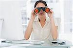 Serious businesswoman looking through binoculars at office desk