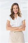 Portrait of a smiling young businesswoman standing with arms crossed against blinds in office