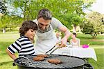 Man and son barbecuing with family in the background at park