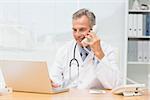 Doctor using laptop and talking on phone at desk in his office at the hospital