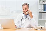 Smiling doctor using laptop and talking on phone at desk in his office at the hospital