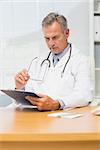 Concentrating mature doctor sitting at his desk with clipboard in his office at the hospital