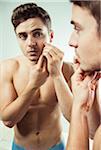 Close-up of young man looking at reflection in bathroom mirror and examining skin on face, studio shot on white background