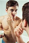 Close-up of young man reflected in bathroom mirror, holding jar of cream and applying to face, studio shot