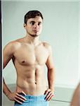 Portrait of young man looking at reflection in bathroom mirror with hands on hips, studio shot