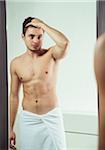 Portrait of young man with towel wrapped around waist, looking in bathroom mirror, studio shot