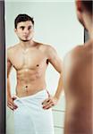 Portrait of young man with towel wrapped around waist, looking in bathroom mirror, studio shot