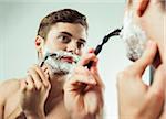 Close-up of Young man looking in bathroom mirror, shaving with razor, studio shot on white background