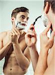 Young man looking in bathroom mirror, shaving with razor, studio shot on white background