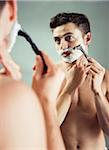 Young man looking in bathroom mirror, shaving with razor, studio shot on grey background