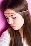 Close-up portrait of young woman with long, brown hair wearing headband, looking downwards, studio shot on pink background