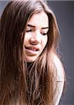 Close-up portrait of young woman with long brown hair, smiling and looking downwards, studio shot on grey background