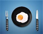 top view of  fried egg on black Plate between silver knife and fork on orange background.