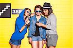 handsome boy in hat shows his cellphone to two beautiful young girls in sunglasses in front of yellow brick wall
