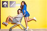 beautiful long haired girl in jeans mini skirt rides a boy in hat on shopping trolley in front of yellow brick wall