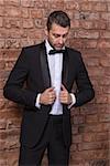 Elegant handsome macho man in a bow tie and tuxedo leaning against a brick wall giving the camera a sultry look