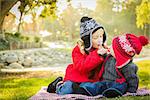 Little Girl with Her Baby Brother Wearing Winter Coats and Hats Sharing a Lollipop Outdoors at the Park.