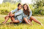 two girlfriends in jeans wear sitting on grass looking at camera