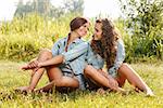 two girlfriends in jeans wear sitting on grass looking at each other