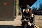 Teenage girl sitting on curb of sidewalk outdoors, wearing fedora and using tablet computer, Germany