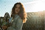 Portrait of teenage girl standing outdoors next to chain link fence, holding skateboard, Germany