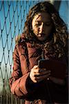 Close-up of teenage girl standing next to chain link fence, wearing winter coat and using smart phone, Germany