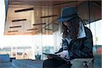 Teenage girl sitting outdoors at train station, wearing fedora and looking at tablet computer, Mannheim, Germany